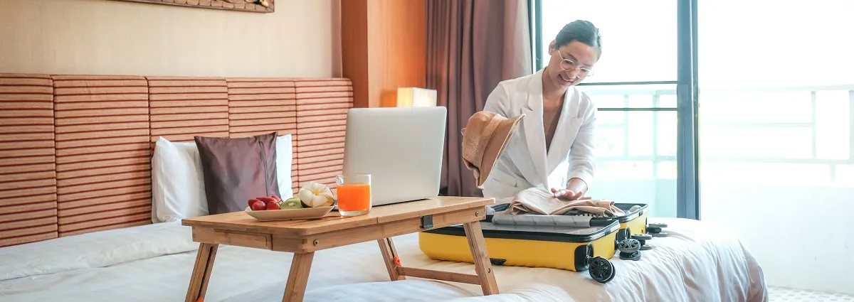 A female hotel guest sits on a bed in a hotel room besides her luggage and a breakfast tray with a plate of fruit, a drink and an open laptop.