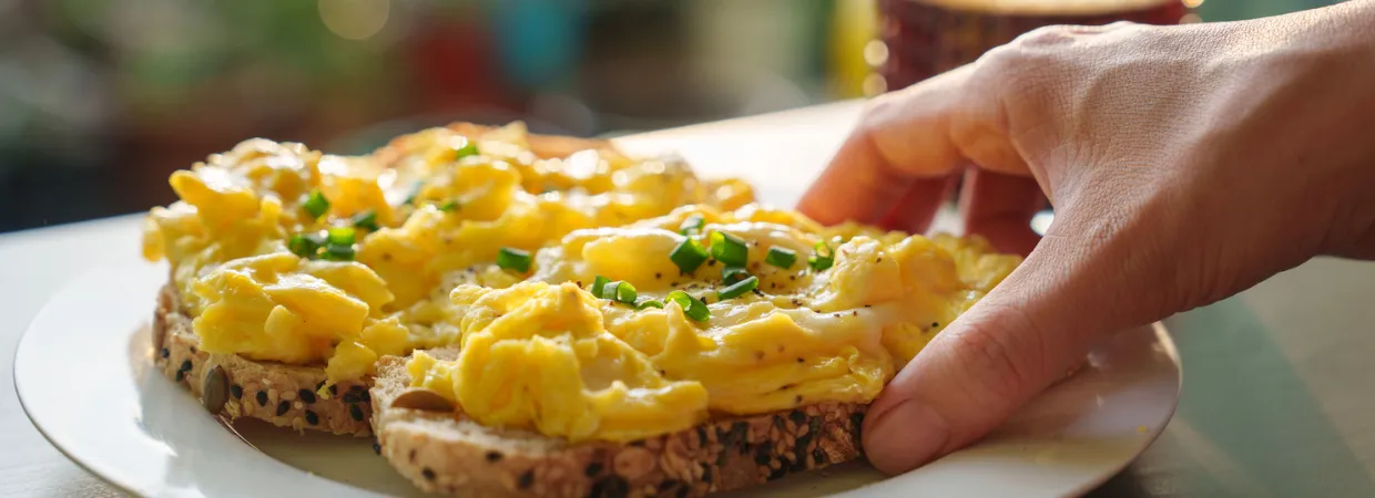Hand reaching for scrambled eggs on multigrain toast, garnished with chives, presenting a fresh breakfast option for hotels, restaurants, and senior living menus.