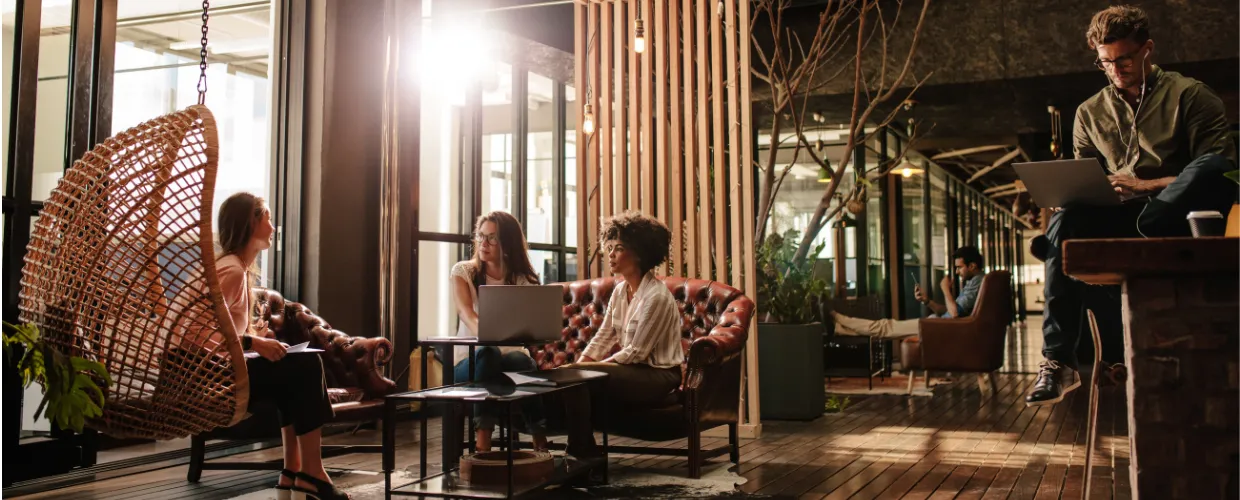Three Gen Z women are seated in a contemporary hotel lobby, two on a leather couch with a laptop, and one in a hanging chair ; a  young man sits on a counter looking at his laptop and another man sits in a lobby lounge chair in the background.